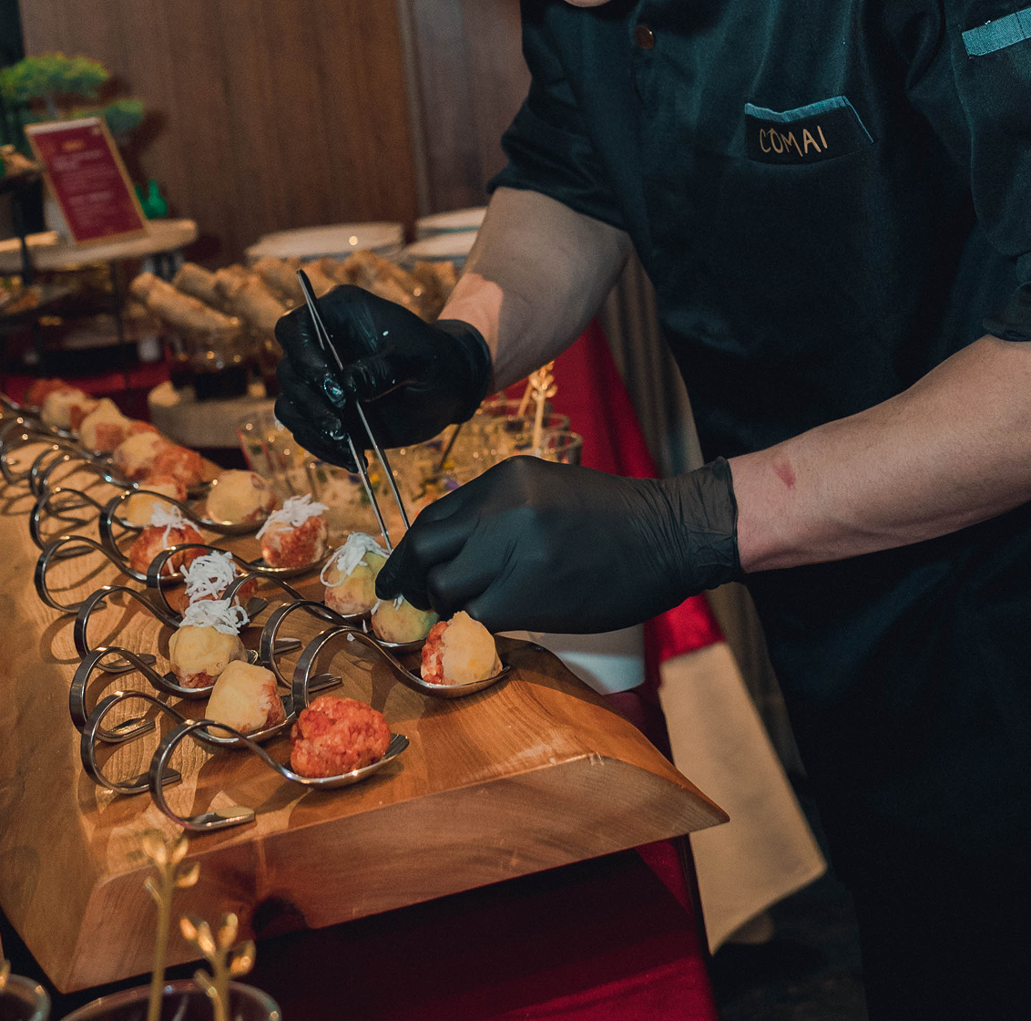 Ein Kellner in schwarzem Outfit richtet Fingerfood auf Löffeln an einem Holzteller an.