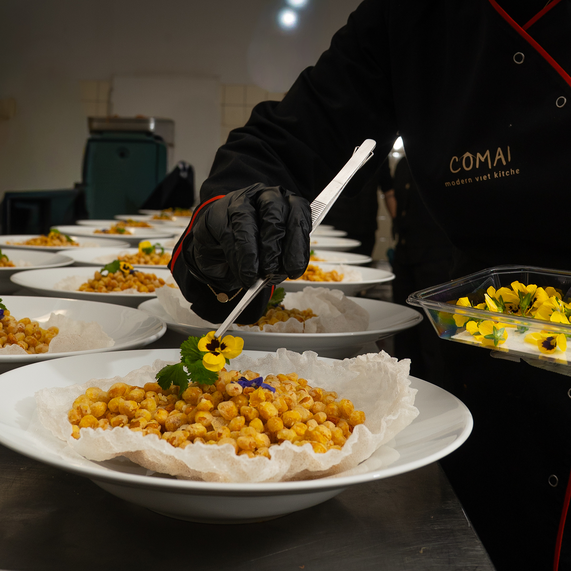Ein Koch dekoriert Teller mit einer goldenen Speise und essbaren Blumen in einer gehobenen Küche.