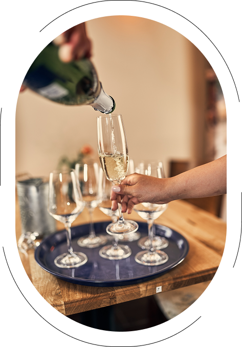 Eine Hand gießt Sekt in ein Glas, während mehrere Gläser auf einem blauen Tablett stehen.