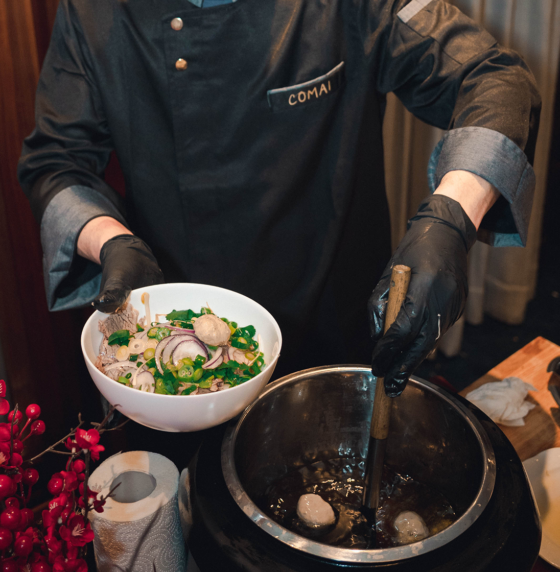 Ein Koch mit Handschuhen serviert eine Schüssel mit frischen Zutaten und würzt eine Suppe aus einem großen Topf.